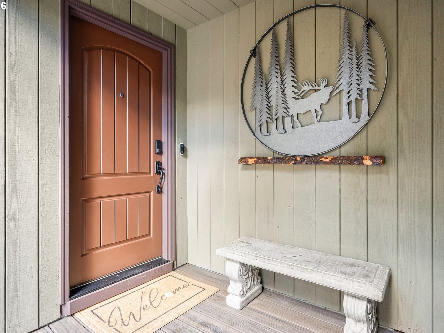 Front entry of 4330 N Shasta Loop with a heavy stained wood door beside a circular wrought-iron elk-in-the-firs sculpture mounted on green board-and-batten siding