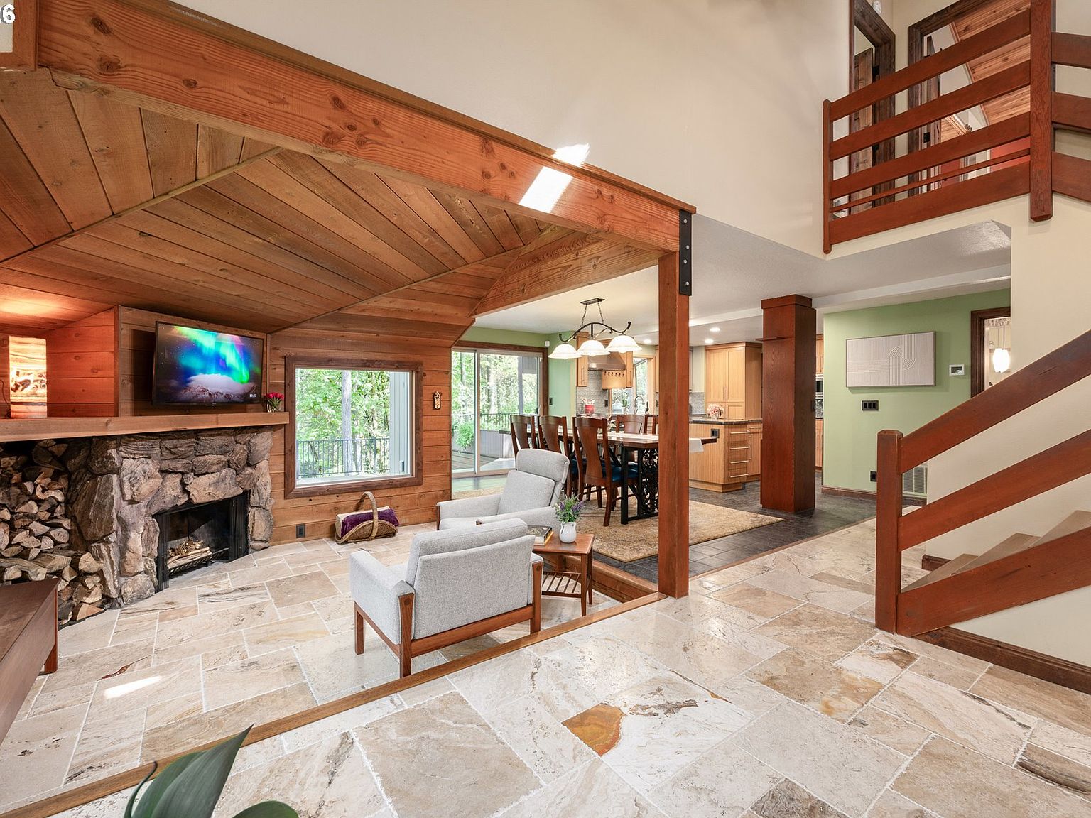 Living and dining areas of the great room from a deeper angle, showing the river-stone fireplace, wood-clad vaults, and dining table sited beside the back window wall